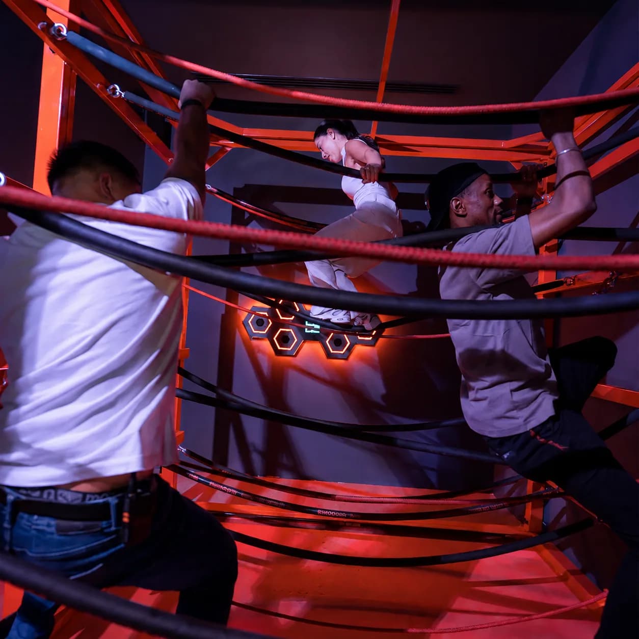 Three colleagues climbing together through a tangle of bungee cords in a Physical-category challenge room during a corporate team-building event